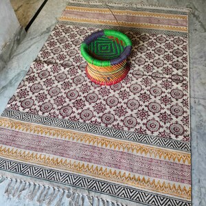 May include: A white and cream patterned rug with a black, yellow, and red border. A woven green, black, and red stool sits on the rug.