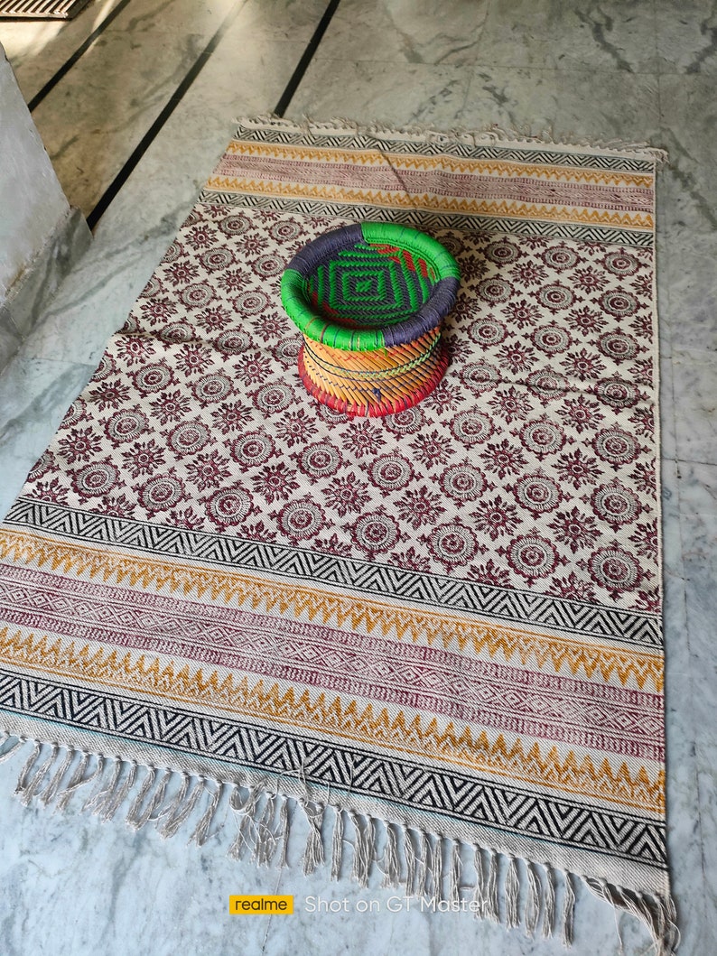 May include: A white and cream patterned rug with a black, yellow, and red border. A woven green, black, and red stool sits on the rug.