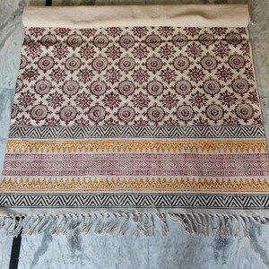 May include: A white cotton rug with a red and black geometric pattern. The rug has a fringe border and is rolled up.
