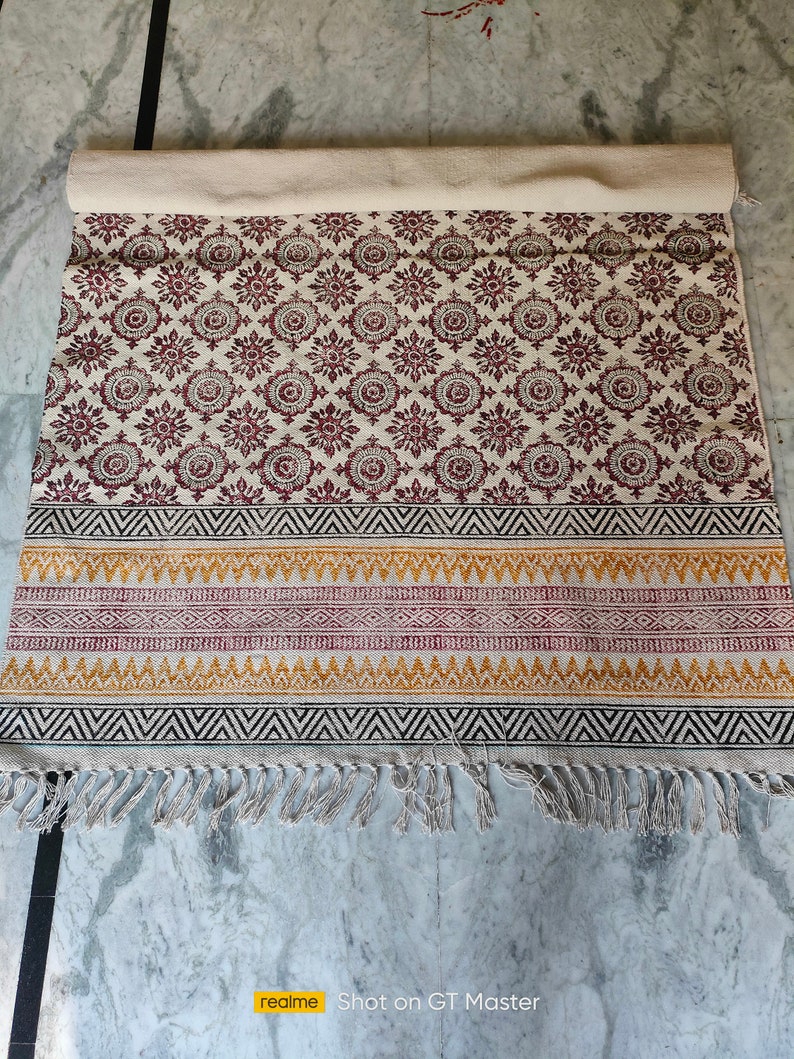 May include: A white cotton rug with a red and black geometric pattern. The rug has a fringe border and is rolled up.
