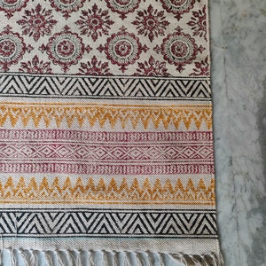 May include: A white cotton rug with a geometric pattern in black, red, and yellow. The rug has a fringe border and is made of natural fibers.