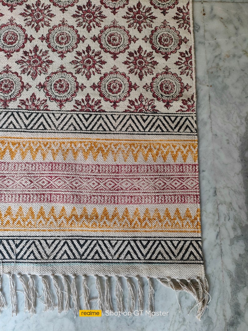 May include: A white cotton rug with a geometric pattern in black, red, and yellow. The rug has a fringe border and is made of natural fibers.