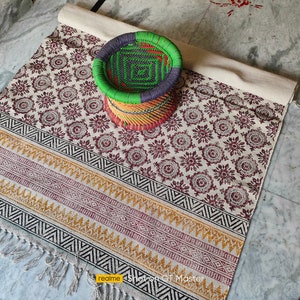 May include: A white and brown patterned rug with black and yellow geometric borders. The rug has fringe on the edges. A woven green, blue, and yellow stool sits on the rug.