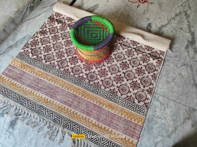 May include: A white and brown patterned rug with black and yellow geometric borders. The rug has fringe on the edges. A woven green, blue, and yellow stool sits on the rug.