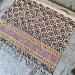 May include: A white cotton rug with a burgundy floral pattern and black, yellow, and burgundy geometric border. The rug has fringe on all sides.