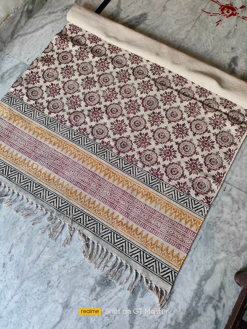May include: A white cotton rug with a burgundy floral pattern and black, yellow, and burgundy geometric border. The rug has fringe on all sides.