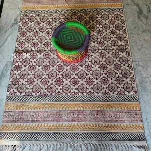May include: A white and brown patterned rug with a woven basket in the center. The basket is green, blue, and red. The rug has a fringe border.