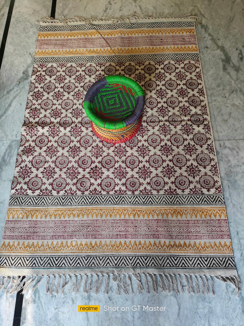 May include: A white and brown patterned rug with a woven basket in the center. The basket is green, blue, and red. The rug has a fringe border.