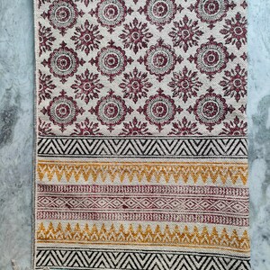 May include: A white cotton rug with a burgundy floral pattern and a black, white, and yellow geometric border. The rug has a fringe on the bottom edge.