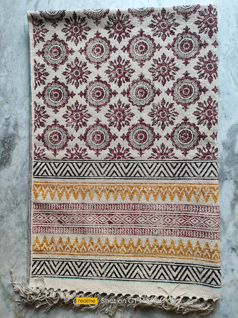 May include: A white cotton rug with a burgundy floral pattern and a black, white, and yellow geometric border. The rug has a fringe on the bottom edge.