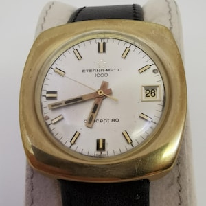Puede incluir: Un reloj de pulsera dorado con una esfera cuadrada y una correa de cuero negra. La esfera del reloj es blanca con marcas negras y el texto "Eterna-Matic 1000" y "concept 80". La ventana de la fecha muestra el número 28.
