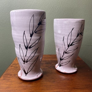May include: Two ceramic mugs with a white glaze and black leaf designs. The mugs are different sizes and have a slightly textured surface.