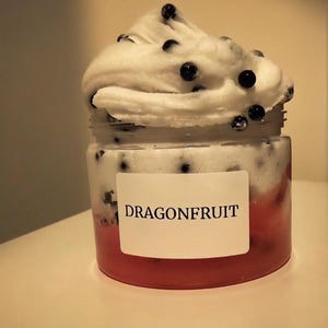 May include: A jar of dragonfruit-themed product with a white, fluffy topping dotted with dark berries. The jar has a red base, a white middle section with black specks, and a label that reads "DRAGONFRUIT".