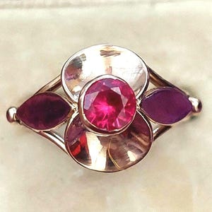 May include: A gold-coloured ring with a central, faceted, round, bright pink gemstone, flanked by two petal-shaped gemstones in a deep magenta hue. The ring's design includes two circular elements on either side of the central stone.