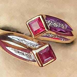 May include: Two gold rings with a bypass design. Each ring features a square, deep red gemstone set in a gold bezel. The rings have a purple band with small, sparkling white stones.