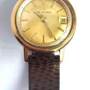 May include: A gold-toned Eternamatic wristwatch with a brown leather band. The watch face is gold with gold hands and markers, and a date window at the 3 o'clock position. The band is a dark brown, textured leather.