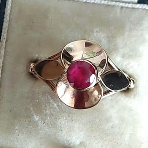 以下が含まれることがあります： 中央に丸い深紅色の宝石をあしらった、金色のリング。リングは、3つの円形と2つの葉の形をした要素を持つ花のモチーフが特徴です。リングは明るい背景にセットされています。