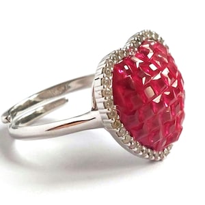 May include: A silver ring with a heart-shaped, ruby-red gemstone. The heart is framed by a halo of small, clear stones. The adjustable band has a split design. The ring is a piece of jewellery.