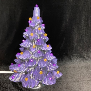 Op de afbeelding: Een keramische kerstboom in een levendige paarse tint, versierd met kleine, ronde, gouden en paarse lampjes. De boom heeft een glanzende afwerking en staat tegen een donkere achtergrond. Een wit netsnoer komt uit de basis.