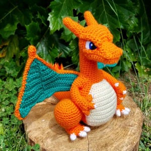 Könnte beinhalten: Handgefertigter, orangefarbener Häkchendrache mit türkisfarbenen Flügeln und weißen Akzenten. Der Drache sitzt auf einem Holzstumpf vor einem Hintergrund aus grünem Laub. Der Drache hat blaue Augen und weiße Krallen.