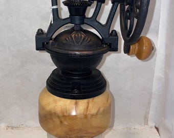 Puede incluir: Un molinillo de café de hierro fundido negro con una manivela de madera y una base de madera. El molinillo tiene un frasco de vidrio para recoger el café molido.