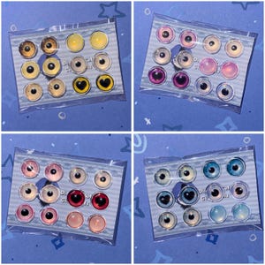 Op de afbeelding: Vier sets poppenogen in verschillende kleuren, waaronder geel, bruin, roze, rood, blauw en groen. Elke set bevat 12 ogen in een doorzichtige plastic verpakking. Sommige ogen hebben hartvormige pupillen. De tekst "Diyful Dolls" is zichtbaar.