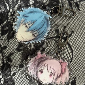 Puede incluir: Dos llaveros de personajes de estilo anime. Un llavero presenta un personaje de pelo azul, y el otro un personaje de pelo rosa. Ambos llaveros tienen cierres plateados y están colocados sobre un fondo de encaje negro.