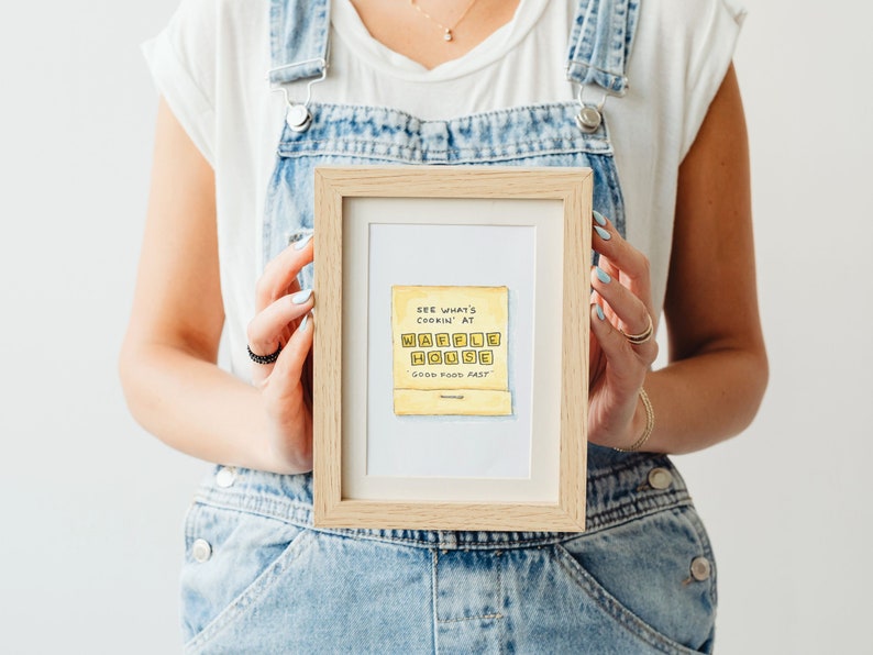 K&ouml;nnte beinhalten: Eine gerahmte Illustration eines gelben Schilds mit dem Text "SEE WHAT'S COOKIN' AT WAFFLE HOUSE GOOD FOOD FAST" in schwarzer Handschrift.