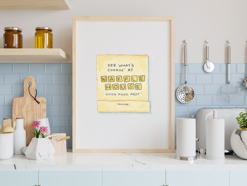 K&ouml;nnte beinhalten: Ein gerahmter Druck einer Aquarellillustration einer gelben Streichholzschachtel mit dem Text "See what's cookin' at Waffle House" und "Good food fast" darauf.