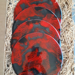 May include: Four round decorative plates with a red and black abstract design, presented in a cardboard box with shredded paper for protection. The plates have a glossy finish and a unique, artistic appearance.
