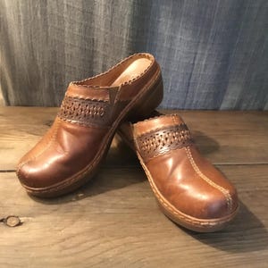 May include: A pair of brown leather clogs featuring a woven cutout detail across the instep, a scalloped collar edge, and a thick sole. The shoes have visible stitching along the toe and sides, with a smooth finish on the leather upper.