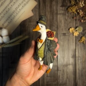 May include: A white goose figurine wearing a black top hat and a grey pinstripe suit, holding a bouquet of red flowers. The goose is standing on a hand.