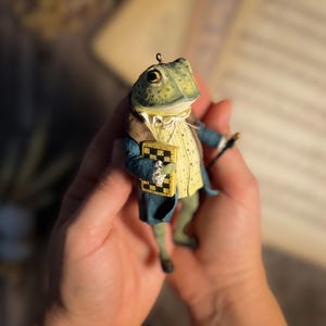 May include: A whimsical frog figurine, dressed in a blue coat, yellow vest, and brown jacket, holding a miniature chessboard and a cane. The frog has a green head and is wearing a white collar. The figurine has a small loop at the top for hanging.