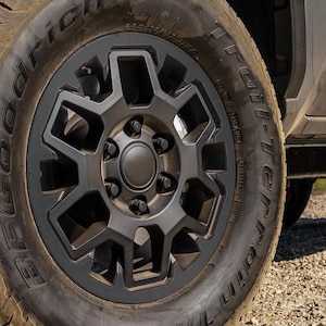 May include: Close-up of a black alloy wheel with a geometric design. The tyre is a BFGoodrich All-Terrain T/A. The wheel is mounted on a vehicle, with a portion of the vehicle's body visible in the background.