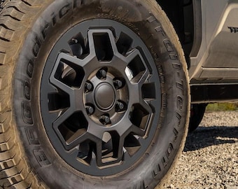 May include: Close-up of a black alloy wheel with a geometric design. The tire is a BFGoodrich All-Terrain T/A. The wheel is mounted on a vehicle, with a portion of the vehicle's body visible in the background.