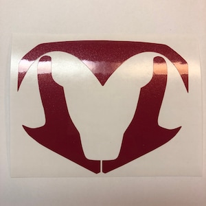 May include: A red glittery decal featuring a stylized ram's head design. The decal is a deep red color and has a shiny, textured appearance. The ram's head is a bold, symmetrical shape.