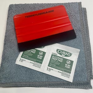 May include: A red plastic squeegee with a black felt edge and the text TVDVINYLDECALS.COM, placed on a gray microfiber cloth alongside two white Curad alcohol prep pad packets, each labeled with green text and a medium prep pad designation.