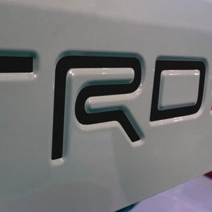 May include: Close-up of a light grey surface with the black letters "TRD" and the red and white letters "PRO" embossed. The letters have a glossy finish and are slightly raised from the surface.