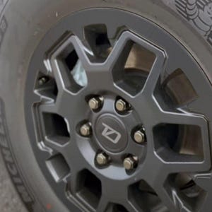 May include: A close-up of a dark grey alloy wheel with a geometric design. The wheel has a central hubcap with a stylized logo and is mounted on a black tyre. The tyre has the Michelin brand name.
