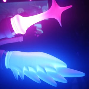 Op de afbeelding: Een futuristische kunstinstallatie met twee verlichte elementen. De ene is een stervorm in roze, de andere een handachtige vorm in blauw. De achtergrond is donker met hints van paars en blauw.