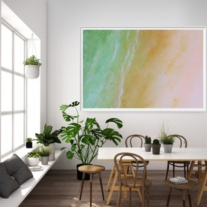 May include: A framed abstract painting of a beach scene with green and blue water and a yellow and pink sandy beach. The painting is hanging on a white wall in a room with a white table and chairs.