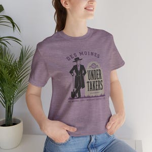May include: A heathered purple short-sleeve t-shirt featuring a graphic of a person in a suit and hat holding a shovel next to a tombstone. The text reads DES MOINES UNDER TAKERS, DES MOINES, IOWA, WESTERN LEAGUE, 1905 in dark purple ink.