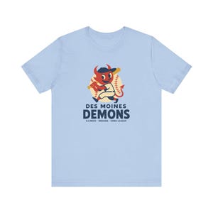 May include: A light blue t-shirt featuring a graphic of a red devil character wearing a baseball cap and holding a bat in front of a baseball. The text reads Des Moines Demons and Illinois-Indiana-Iowa League in dark blue lettering.