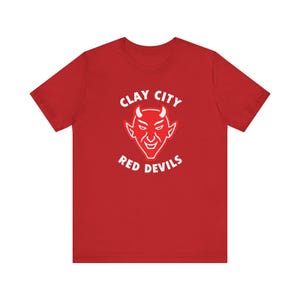 May include: A red short-sleeved t-shirt featuring a white graphic of a devil face centred on the chest. Above the devil graphic is the text CLAY CITY and below the graphic is the text RED DEVILS, both printed in a bold white sans-serif font.