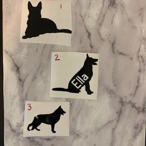 Könnte beinhalten: Drei schwarze Aufkleber mit Silhouette eines Deutschen Schäferhundes auf weißen Quadraten. Ein Aufkleber trägt den Namen "Ella". Die Aufkleber sind mit roter Tinte mit 1, 2 und 3 nummeriert. Der Hintergrund ist ein Marmormuster.