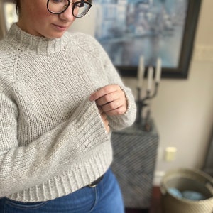 May include: A light gray knitted sweater with a round neckline and long sleeves. The sweater is made of a soft, textured yarn.