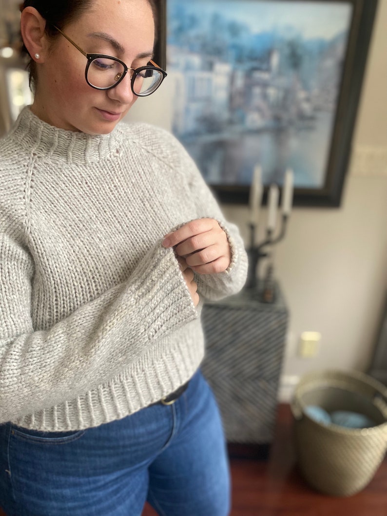 May include: A light gray knitted sweater with a round neckline and long sleeves. The sweater is made of a soft, textured yarn.
