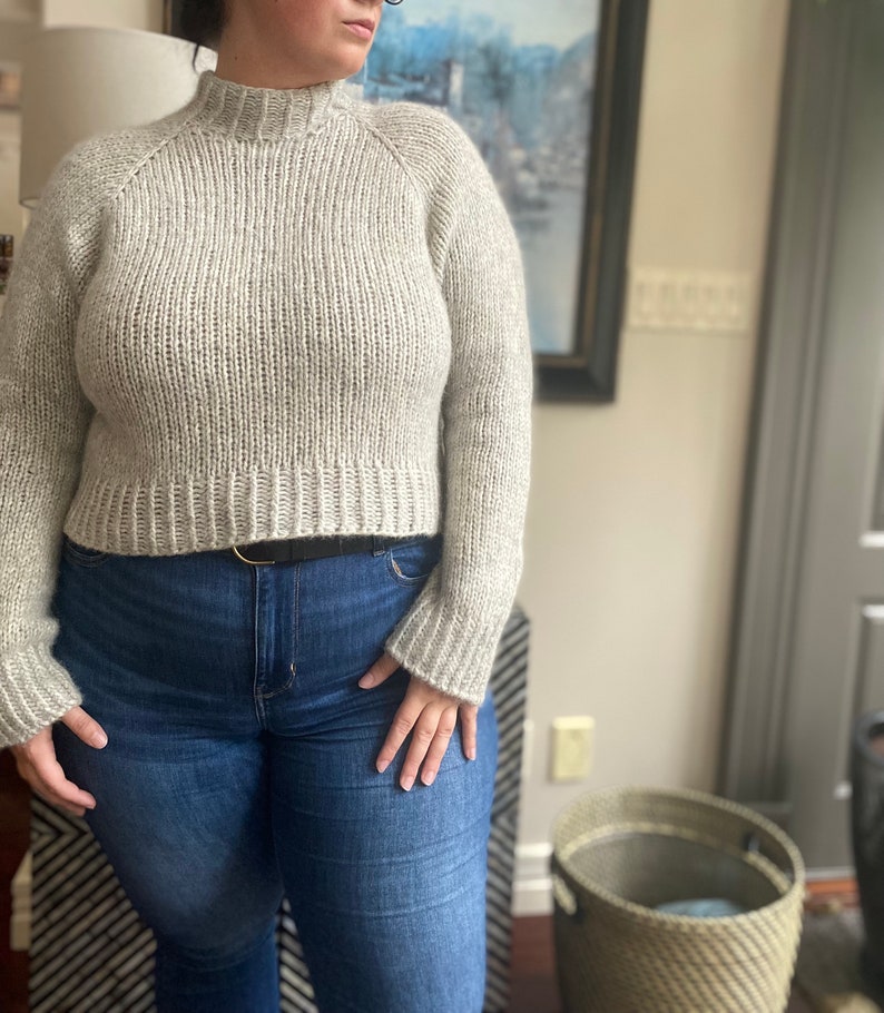May include: A light gray cropped sweater with a mock neck and long sleeves. The sweater is made of a soft, textured yarn and has a relaxed fit.