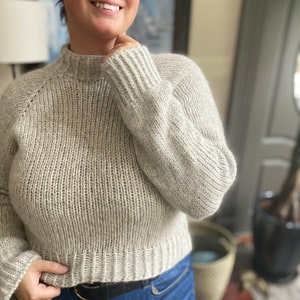 May include: A light gray knitted sweater with a mock neck and long sleeves. The sweater is cropped and has a ribbed texture. The model is wearing the sweater with blue jeans and a black belt.