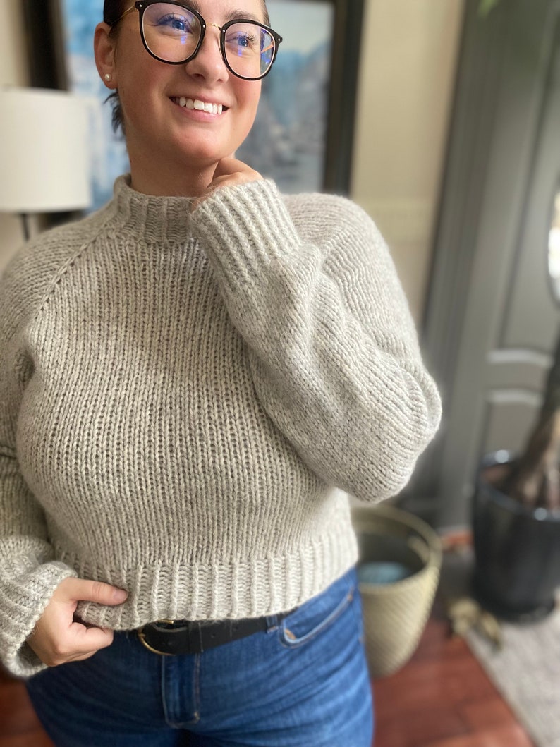 May include: A light gray knitted sweater with a mock neck and long sleeves. The sweater is cropped and has a ribbed texture. The model is wearing the sweater with blue jeans and a black belt.